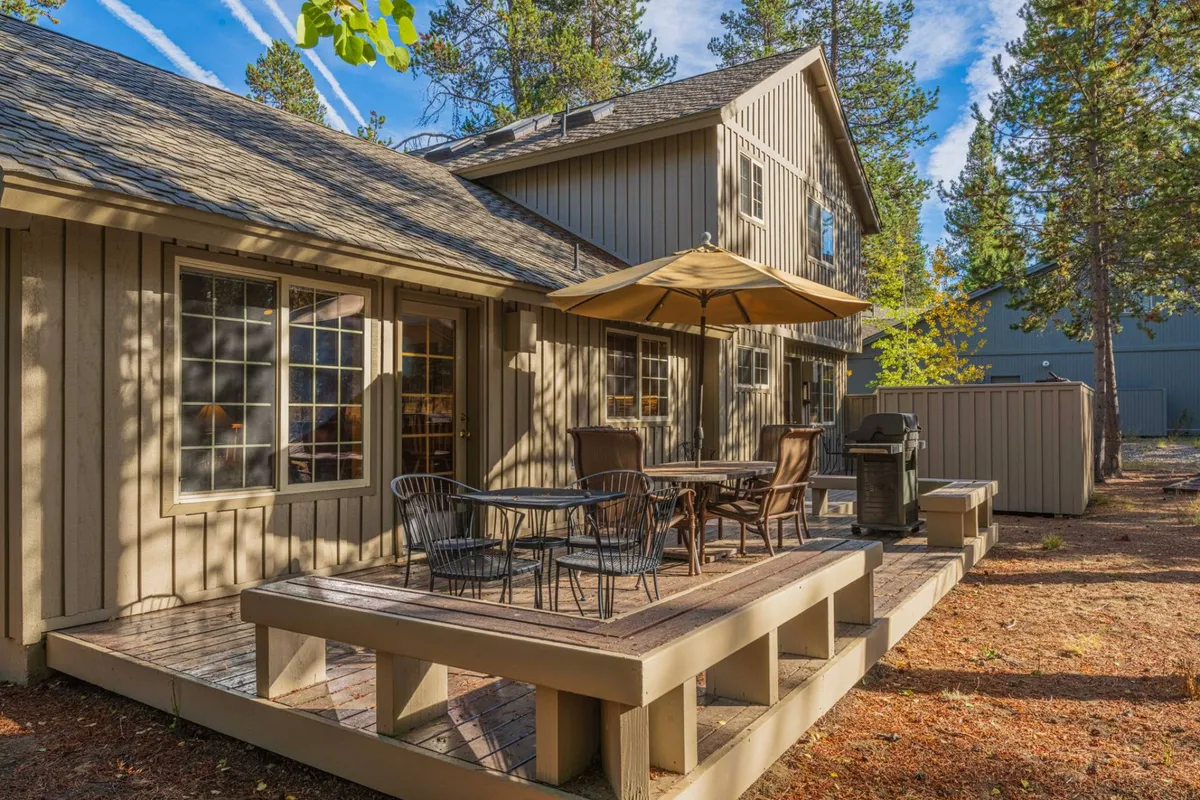 This image shows a welcoming outdoor deck with comfortable seating, an outdoor grill, an umbrella for shade, and the wooden exterior of the house. The space is surrounded by trees, creating a private and serene setting for gatherings.
