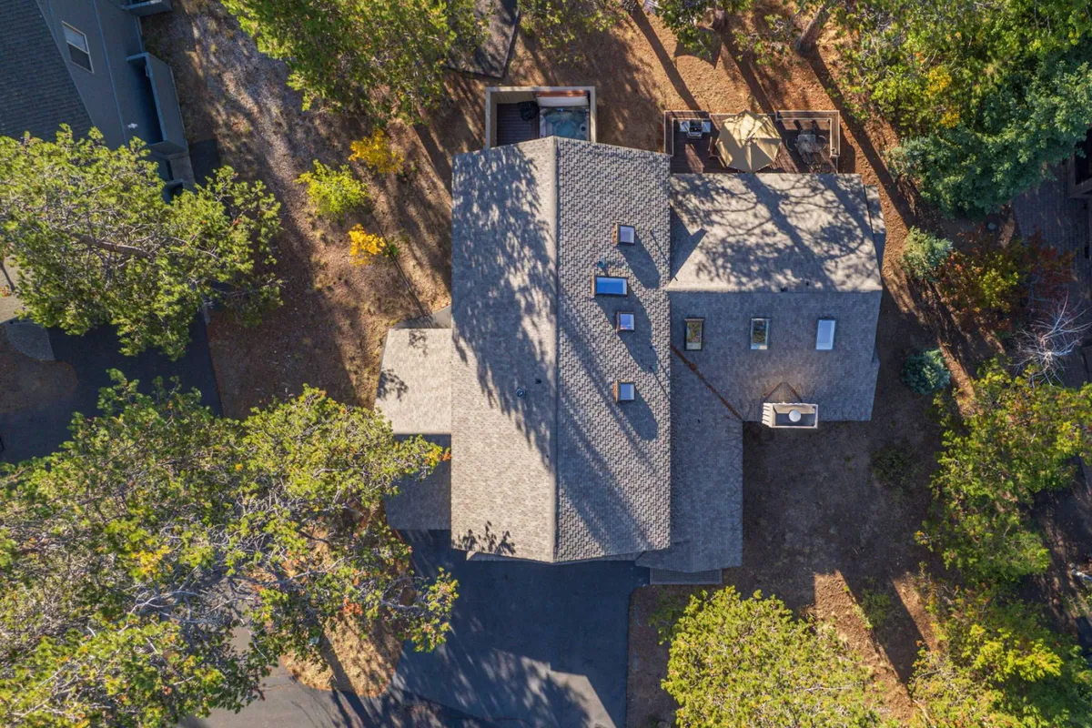 This image captures the exterior of the home, highlighting its distinctive roof design, skylights, and a deck area nestled among tall, lush trees. The scene is tranquil and suggests a seamless connection to nature, with the inviting deck positioned to enjoy sunlight and forest views.