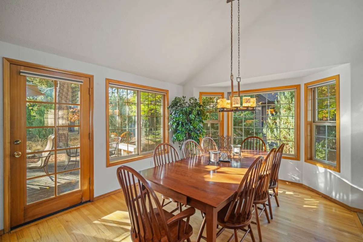 A bright dining area featuring a sturdy dining table, surrounded by large windows that showcase woodland scenery. Green plants and a stylish hanging light fixture add to the warm, welcoming atmosphere, making the space perfect for family dinners or festive occasions.