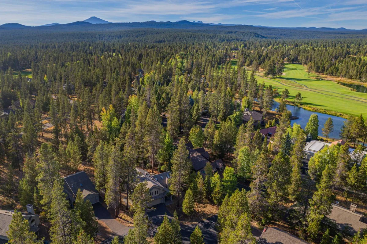 This image displays a sweeping view of a lush golf course surrounded by dense forest, with a tranquil pond and residential homes visible in the background. The mountain range in the distance sets a striking high-desert scene, ideal for illustrating the area's natural beauty and outdoor recreation options.