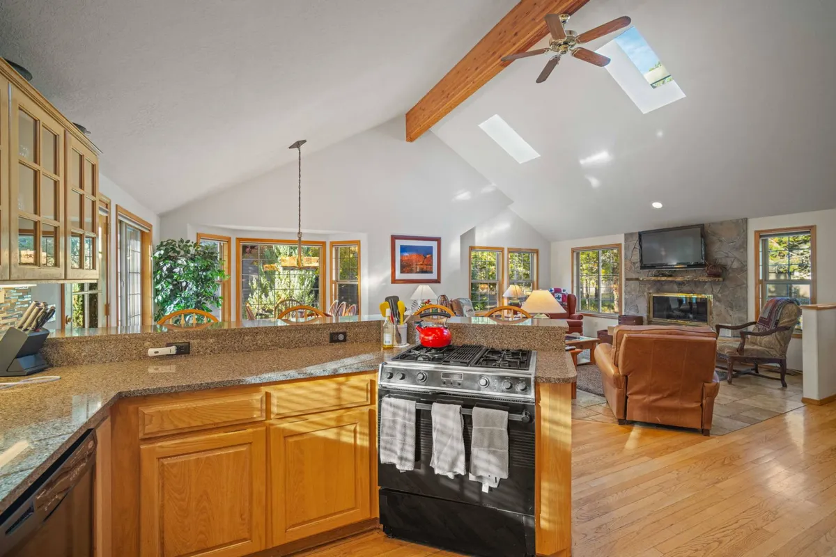 A cozy kitchen scene featuring a granite countertop, stainless steel stove, large bright windows, rustic wooden beams overhead, and a glimpse of a fireplace. The ambiance is warm and inviting, perfect for sharing stories over meals.
