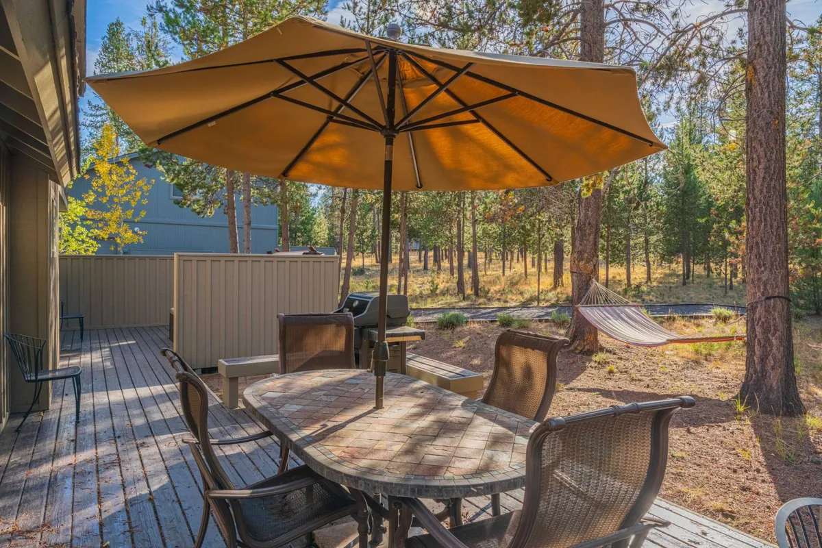 An inviting outdoor dining area sits beneath a large umbrella, with a hammock strung between trees in the background. The scene is enveloped by lush greenery, creating an idyllic spot for al fresco meals, relaxation, or gathering with friends and family just steps from the home.