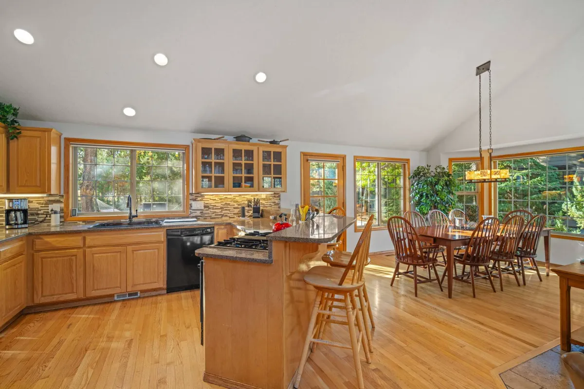 An open-concept kitchen and dining area with a prominent kitchen island, a large wooden dining table set for a group, expansive windows flooding the space with light, and warm wood cabinetry. The kitchen seamlessly flows into the dining area, emphasizing the communal, social atmosphere.