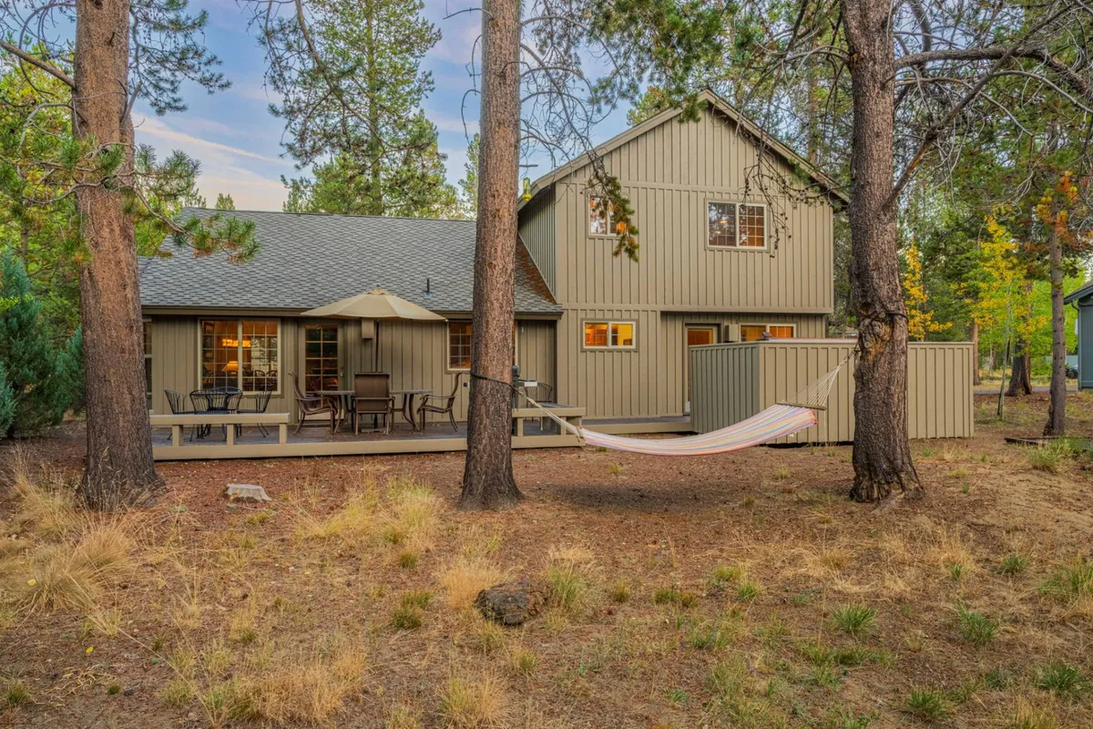 A peaceful backyard setting with a hammock strung between pine trees, a dining set on the deck, and expansive windows that reflect the surrounding nature. The scene suggests both relaxation and outdoor family gatherings.