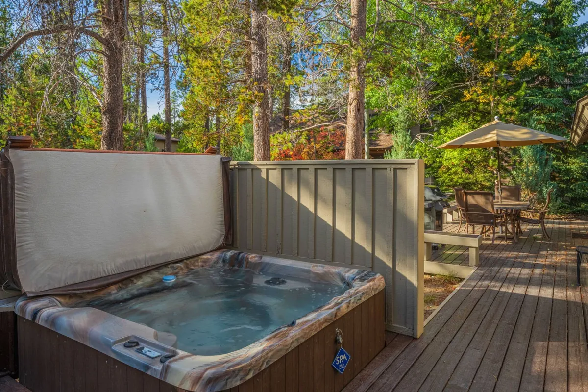 This exterior image features a spacious deck area equipped with a private hot tub and an outdoor dining space, surrounded by lush trees. The scene is tranquil and inviting, with the hot tub positioned for relaxation under the open sky, and the dining area set for family meals or evening gatherings.