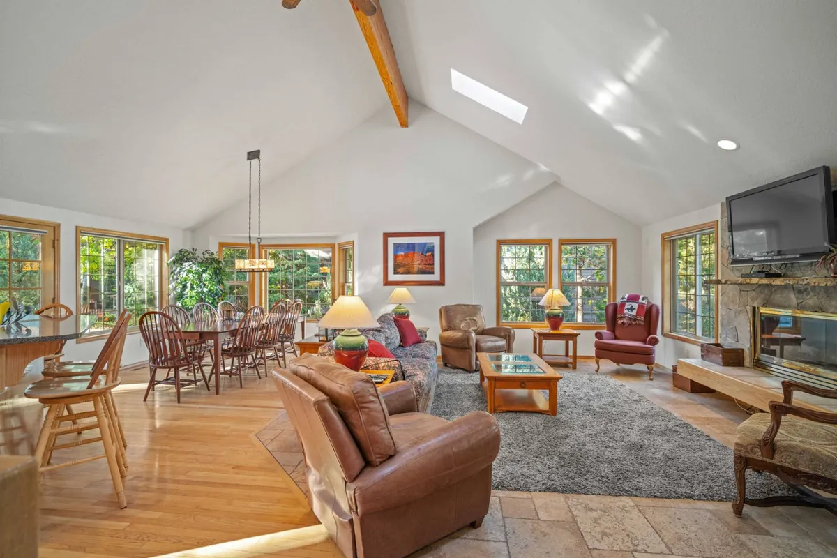 The image showcases a spacious, inviting great room with soaring vaulted ceilings, expansive windows letting in natural light, a grand fireplace as the centerpiece, a large dining table perfect for gatherings, and plush seating arranged for relaxation. Decorative accents and warm tones create a welcoming, family-friendly atmosphere.