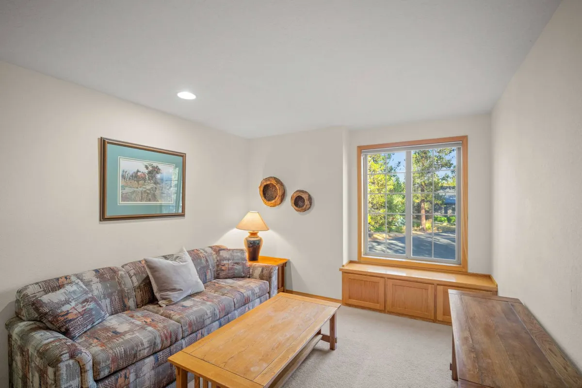 A comfortable living space featuring a sofa, coffee table, window, lamp, and wall art. The room is filled with natural sunlight and exudes a cozy atmosphere, ideal for relaxing, socializing, or converting the space for extra sleeping needs.